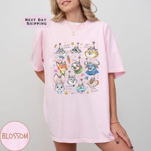 Puede incluir: Camiseta rosa claro con retratos de personajes de dibujos animados con sombreros de fiesta. La camiseta tiene el texto "Next Day Shipping" y "Blossom". El gr&aacute;fico incluye personajes como un zorro, un conejo y un perezoso.