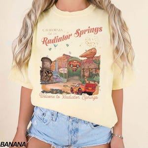 Puede incluir: Camiseta amarillo claro con un dise&ntilde;o de Radiator Springs, con texto que dice "California Est. 2012" y "Radiator Springs". El dise&ntilde;o incluye un coche de dibujos animados y un paisaje des&eacute;rtico.