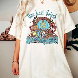 以下が含まれることがあります： クリーム色のTシャツ。カラフルなグラフィックが特徴で、「One Last Splash」の文字と、漫画のキャラクター、そして「Splash Mountain」と「1989 2023」の文字がデザインされています。