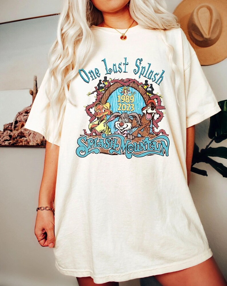 以下が含まれることがあります： クリーム色のTシャツ。カラフルなグラフィックが特徴で、「One Last Splash」の文字と、漫画のキャラクター、そして「Splash Mountain」と「1989 2023」の文字がデザインされています。
