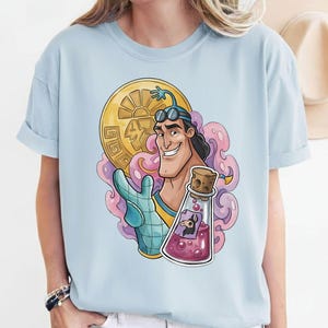 May include: Light blue t-shirt featuring a cartoon illustration of a smiling character holding a gold coin and a potion bottle. The character is wearing goggles and a glove. The coin has a sun design.