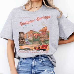 Puede incluir: Camiseta gris claro con un gr&aacute;fico colorido de Radiator Springs, con el texto "California Est. 2011 Radiator Springs" y "Welcome to Radiator Springs". El dise&ntilde;o incluye una torre de agua, un paisaje des&eacute;rtico y dos coches de carreras.