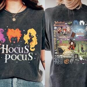 May include: Dark gray t-shirt featuring the text "Hocus Pocus" with illustrations of three witches' hair. The back of the shirt has a collage of images and text related to the movie, including "Winifred Sanderson" and "It's just a bunch of Hocus Pocus."