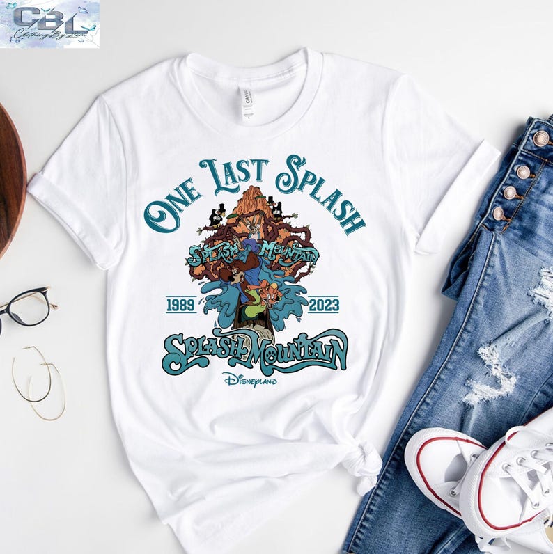 May include: White t-shirt featuring a graphic design for the Splash Mountain ride at Disneyland. The design includes the text "One Last Splash" and "Splash Mountain" with the years 1989 and 2023. The shirt is paired with blue jeans and white sneakers.