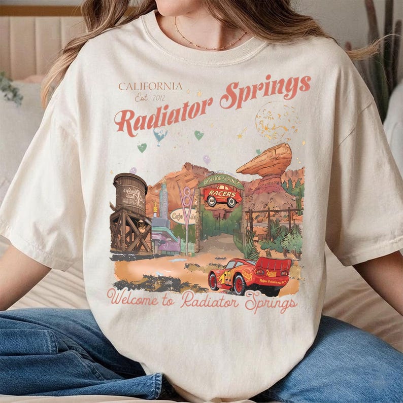 Puede incluir: Camiseta color crema con un dise&ntilde;o de Radiator Springs, con texto que dice "California Est. 2012" y "Radiator Springs". El dise&ntilde;o incluye un coche de carreras rojo y un paisaje des&eacute;rtico. "Welcome to Radiator Springs" est&aacute; impreso en la parte inferior.