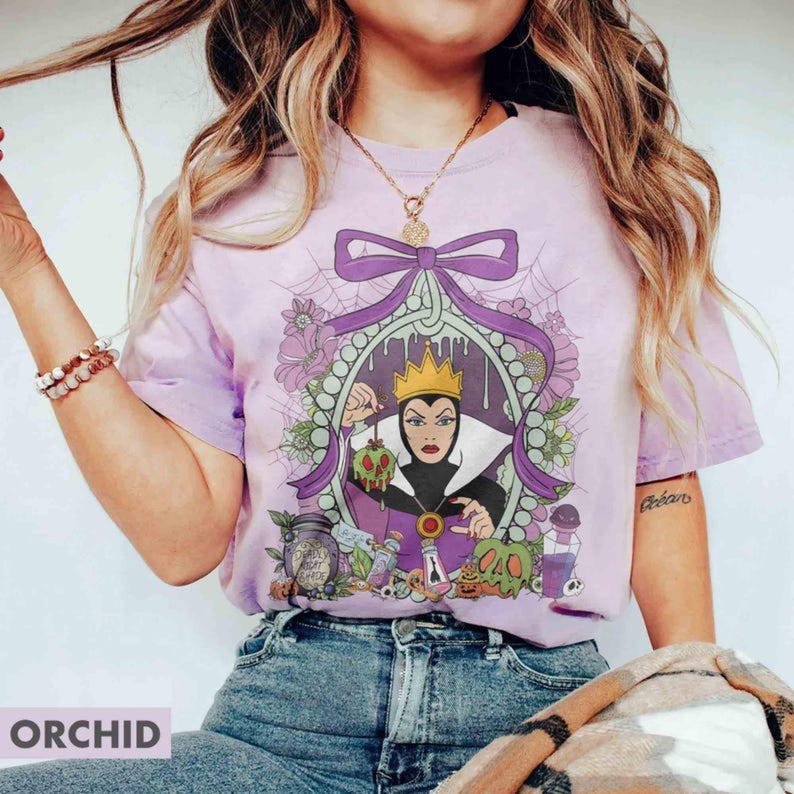 Pu&ograve; includere: T-shirt color orchidea con un'illustrazione a cartoni animati di una regina con una corona, che tiene in mano una mela avvelenata. Il design include un fiocco viola, elementi floreali e la scritta "Evil Night Shade".