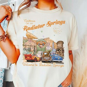 May include: Cream-colored t-shirt featuring a graphic of Radiator Springs, with text that reads "California Est. 2012 Radiator Springs" and "Welcome to Radiator Springs." The design includes cartoon cars, buildings, and desert scenery.