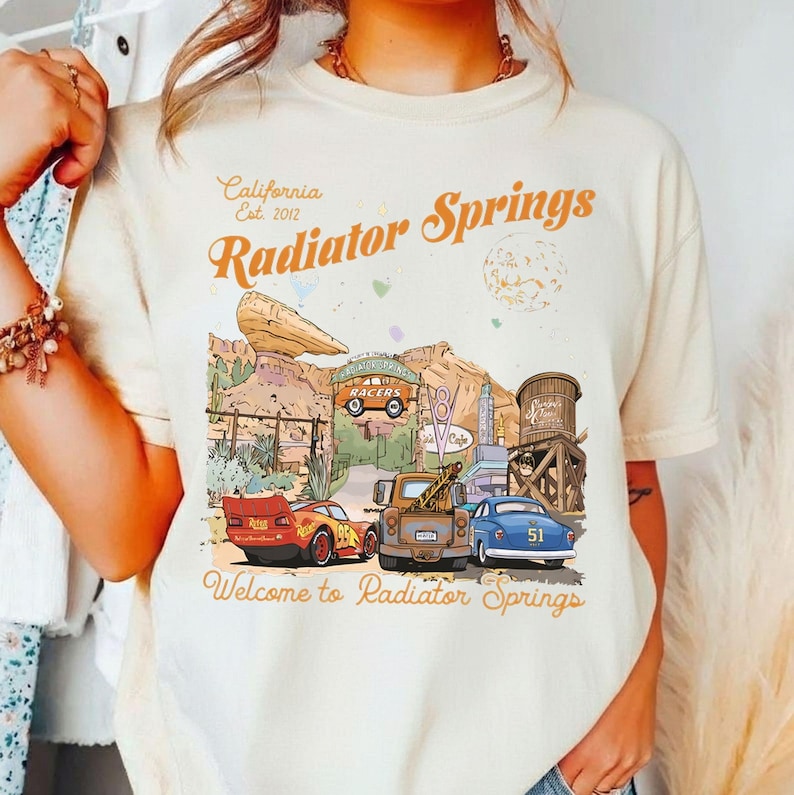 May include: Cream-colored t-shirt featuring a graphic of Radiator Springs, with text that reads "California Est. 2012 Radiator Springs" and "Welcome to Radiator Springs." The design includes cartoon cars, buildings, and desert scenery.