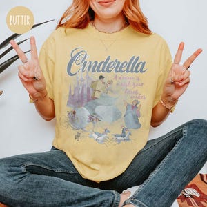 May include: A yellow t-shirt featuring a graphic of Cinderella and Prince Charming. The text on the shirt reads "Cinderella A dream is a wish your heart makes".