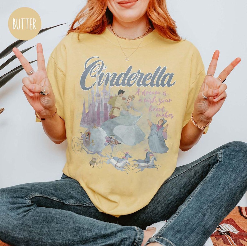 May include: A yellow t-shirt featuring a graphic of Cinderella and Prince Charming. The text on the shirt reads "Cinderella A dream is a wish your heart makes".
