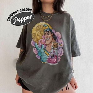 May include: A gray t-shirt featuring a cartoon character with a gold coin, a potion bottle, and the text "COMFORT COLORS Pepper". The graphic is in shades of pink, purple, blue, and yellow. The shirt has short sleeves and a relaxed fit.