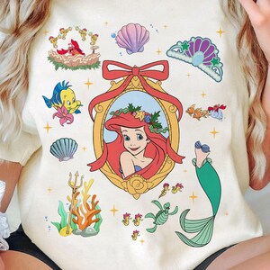 May include: A white t-shirt with a colorful illustration of Ariel, the Little Mermaid, surrounded by seashells, starfish, and other ocean-themed imagery. The illustration includes a red bow, a gold frame, and a green seaweed design.