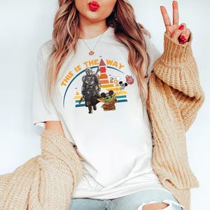 May include: Cream-colored t-shirt featuring a graphic of The Mandalorian and Grogu, with the text "THIS IS THE WAY" in orange and yellow. The design includes a rainbow and Mickey Mouse balloons.