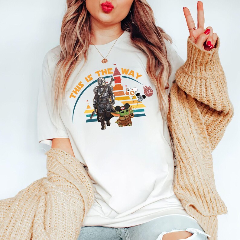 May include: Cream-colored t-shirt featuring a graphic of The Mandalorian and Grogu, with the text "THIS IS THE WAY" in orange and yellow. The design includes a rainbow and Mickey Mouse balloons.