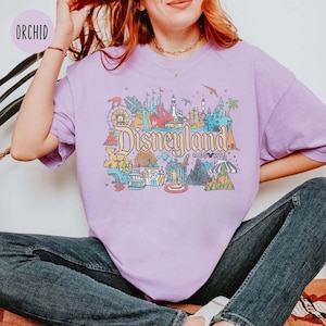 May include: A purple t-shirt with a colorful illustration of Disneyland Resort, including the iconic castle, rides, and other attractions. The text "Disneyland Resort" is printed on the shirt.