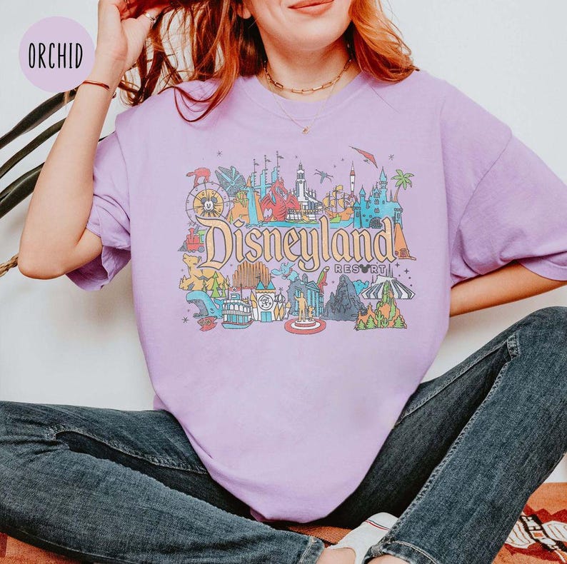May include: A purple t-shirt with a colorful illustration of Disneyland Resort, including the iconic castle, rides, and other attractions. The text "Disneyland Resort" is printed on the shirt.