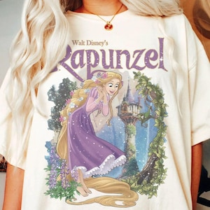 May include: Cream-colored t-shirt featuring the text "Walt Disney's Rapunzel" in purple. The graphic depicts Rapunzel in a purple dress with long blonde hair, a castle, and floral elements. A casual, comfortable tee.