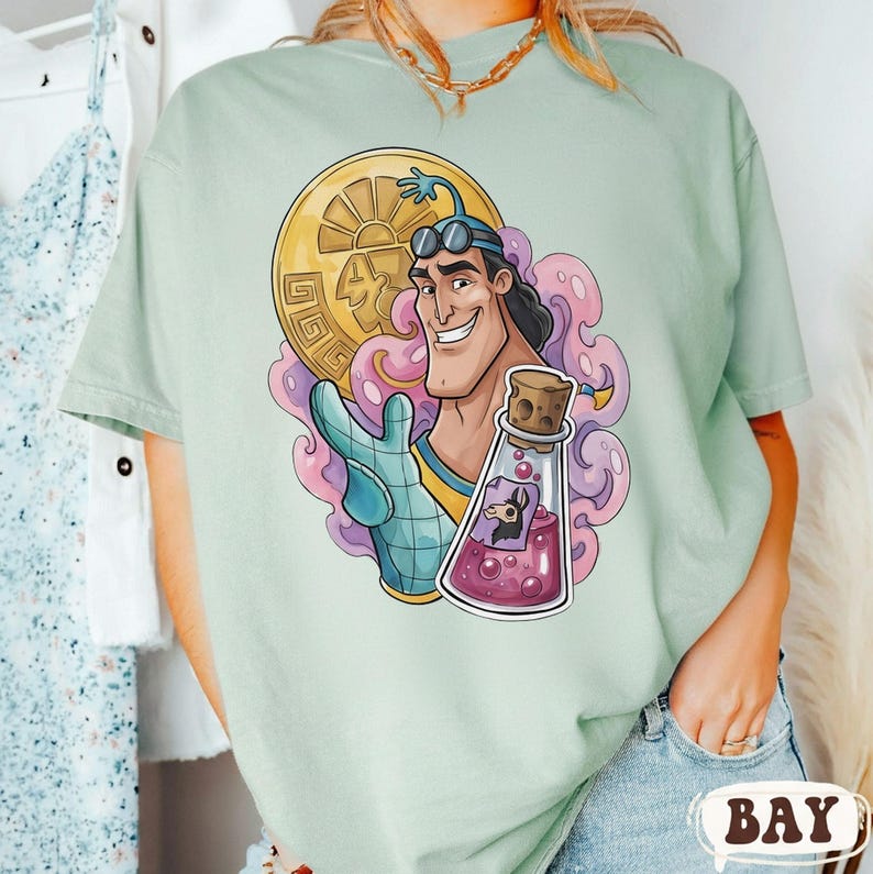 May include: A light green t-shirt featuring a cartoon character holding a gold coin and a potion bottle. The character has a tan complexion, black hair, and is wearing blue goggles. The potion bottle contains a black llama. The design is surrounded by pink and purple swirls.