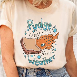 May include: Cream-colored t-shirt featuring a cartoon design. The graphic includes an orange creature with blue spots, a slice of toast, and the text "Pudge Controls the Weather" in blue. The design is whimsical and playful.