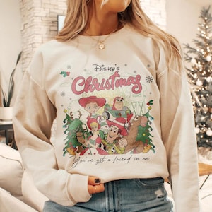Może przedstawiać: Kremowa bluza z nadrukiem postaci z Toy Story Disneya i napisem "Disney's Christmas" w kolorze r&oacute;żowym. Poniżej postaci wydrukowano frazę "You've got a friend in me". Projekt zawiera płatki śniegu i choinki.