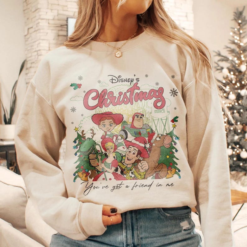Może przedstawiać: Kremowa bluza z nadrukiem postaci z Toy Story Disneya i napisem "Disney's Christmas" w kolorze r&oacute;żowym. Poniżej postaci wydrukowano frazę "You've got a friend in me". Projekt zawiera płatki śniegu i choinki.