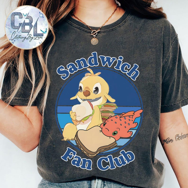 May include: Dark grey t-shirt with a cartoon design. The design features a yellow character holding a sandwich, a red fish-like creature, and the text "Sandwich Fan Club" in blue, set within a circular frame.