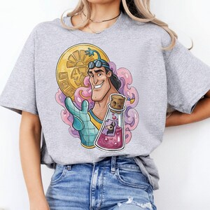 May include: A light gray t-shirt featuring a colorful graphic of a cartoon character holding a gold coin and a potion bottle. The graphic includes pink and purple accents. The shirt is paired with blue jeans.