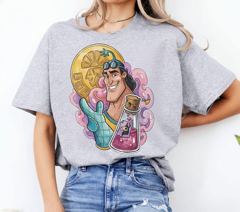 May include: A light gray t-shirt featuring a colorful graphic of a cartoon character holding a gold coin and a potion bottle. The graphic includes pink and purple accents. The shirt is paired with blue jeans.
