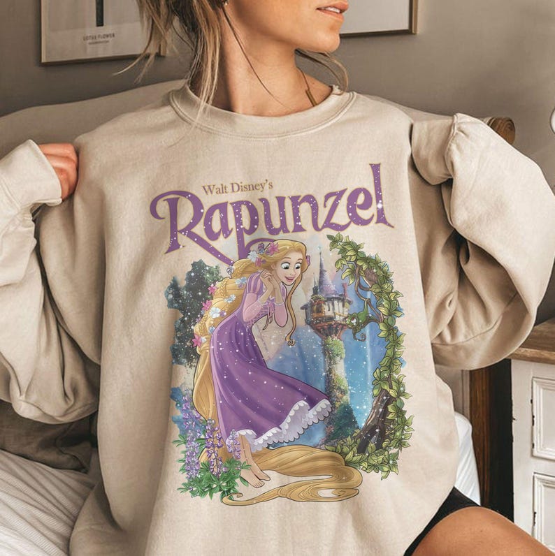May include: Beige sweatshirt featuring a graphic of Rapunzel from Walt Disney's animated film. The illustration shows Rapunzel in a purple dress with long blonde hair, a castle, and the text "Walt Disney's Rapunzel" in purple.