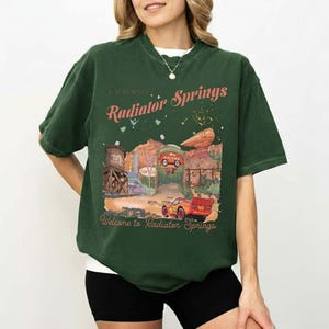 Puede incluir: Camiseta verde oscuro con un gr&aacute;fico de Radiator Springs de la pel&iacute;cula Cars. El dise&ntilde;o incluye un coche de carreras rojo, un paisaje des&eacute;rtico y el texto "Radiator Springs" y "Welcome to Radiator Springs".