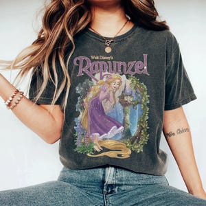 May include: Dark gray t-shirt featuring a graphic of Rapunzel from Walt Disney's animated film. The design includes Rapunzel in a purple dress with long blonde hair, a castle, and floral accents. The text "Walt Disney's Rapunzel" is arched above the image.