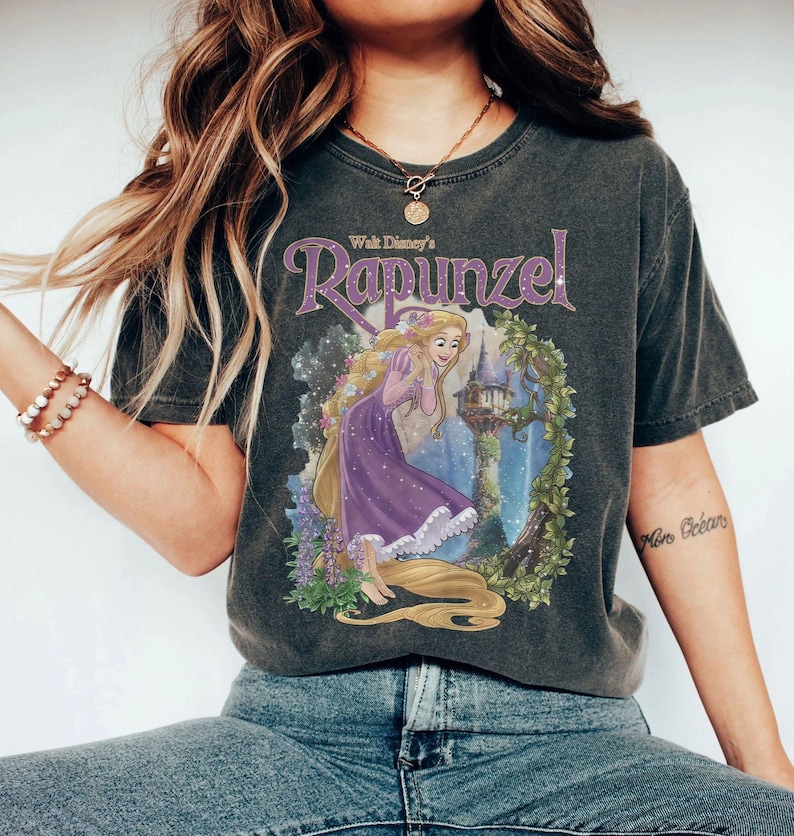 May include: Dark gray t-shirt featuring a graphic of Rapunzel from Walt Disney's animated film. The design includes Rapunzel in a purple dress with long blonde hair, a castle, and floral accents. The text "Walt Disney's Rapunzel" is arched above the image.