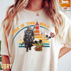 May include: Ivory-colored t-shirt featuring a graphic design with the text "THIS IS THE WAY." The design includes illustrations of The Mandalorian, Grogu, and themed balloons. The shirt has a crew neck and short sleeves.