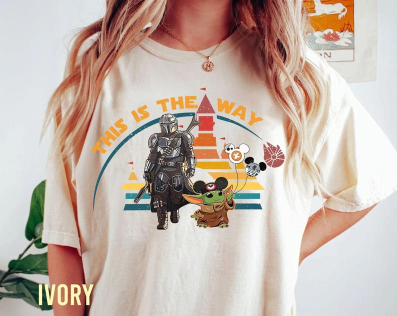 May include: Ivory-colored t-shirt featuring a graphic design with the text "THIS IS THE WAY." The design includes illustrations of The Mandalorian, Grogu, and themed balloons. The shirt has a crew neck and short sleeves.