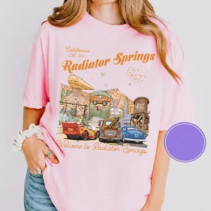 May include: Pink t-shirt featuring a graphic of Radiator Springs from the movie Cars. The design includes the text "California Est. 2012" and "Welcome to Radiator Springs". The graphic shows various cars and buildings.