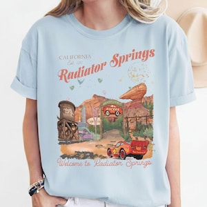 Puede incluir: Camiseta azul claro con un dise&ntilde;o de Radiator Springs, con texto que dice "California Est. 2012 Radiator Springs" y "Welcome to Radiator Springs". El dise&ntilde;o incluye un coche de dibujos animados y un paisaje des&eacute;rtico.