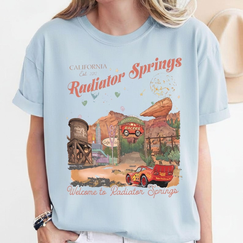 Puede incluir: Camiseta azul claro con un dise&ntilde;o de Radiator Springs, con texto que dice "California Est. 2012 Radiator Springs" y "Welcome to Radiator Springs". El dise&ntilde;o incluye un coche de dibujos animados y un paisaje des&eacute;rtico.