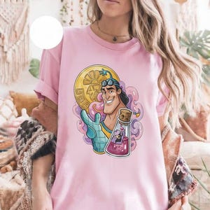 May include: Pink t-shirt featuring a cartoon character with a gold coin, a potion bottle, and a blue glove. The character has black hair and goggles. The potion bottle contains a purple liquid and a small figure.