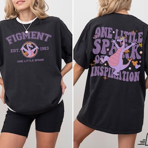 Op de afbeelding: Zwart T-shirt met een paarse draak afbeelding en de tekst "One Little Spark of Inspiration". De voorkant van het shirt heeft een paarse draak afbeelding en de tekst "Figment Est. 1983 One Little Spark".