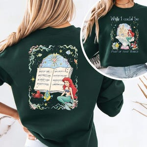 May include: Dark green sweatshirt featuring a colorful illustration of a mermaid with red hair and a turquoise tail, sitting near an open book of sheet music. The design includes sea creatures and the text "Wish I could be Part of That World."