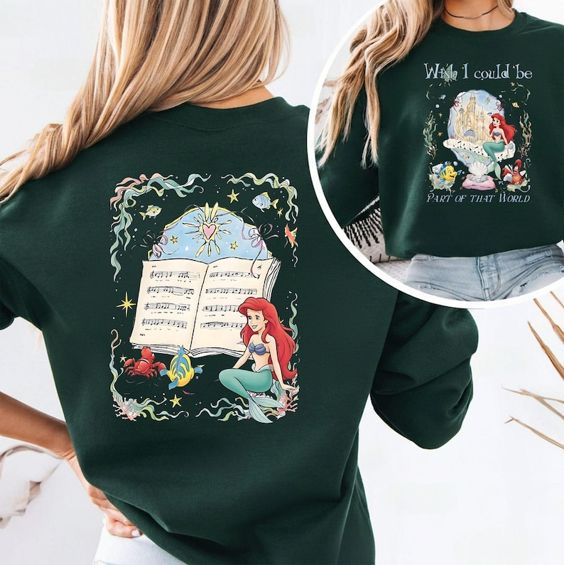 May include: Dark green sweatshirt featuring a colorful illustration of a mermaid with red hair and a turquoise tail, sitting near an open book of sheet music. The design includes sea creatures and the text "Wish I could be Part of That World."