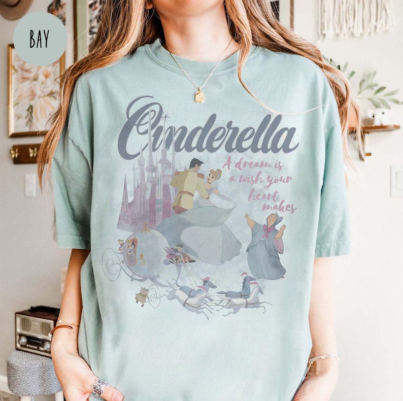 May include: A light blue t-shirt featuring a graphic print of Cinderella and Prince Charming in a carriage. The text "Cinderella" is printed in a large, stylised font above the image. The text "A dream is a wish your heart makes" is printed below the image.