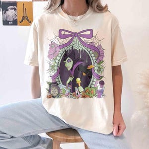 Pu&ograve; includere: T-shirt color crema con grafica di un ritratto con cornice viola di una donna dalla pelle verde e un corvo nero. Il design include fiori viola, ragnatele e bottiglie di pozioni. Il testo "Deadly Night Shade" &egrave; visibile su un barattolo.