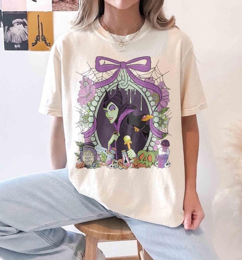 Pu&ograve; includere: T-shirt color crema con grafica di un ritratto con cornice viola di una donna dalla pelle verde e un corvo nero. Il design include fiori viola, ragnatele e bottiglie di pozioni. Il testo "Deadly Night Shade" &egrave; visibile su un barattolo.