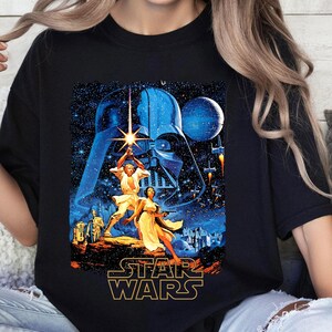 Könnte beinhalten: Ein schwarzes T-Shirt mit einem Vintage-Filmposter-Design von Star Wars, das Darth Vader, Luke Skywalker, Prinzessin Leia und R2-D2 zeigt. Das Poster ist vor einem dunkelblauen Weltraumhintergrund mit Sternen und Planeten platziert.
