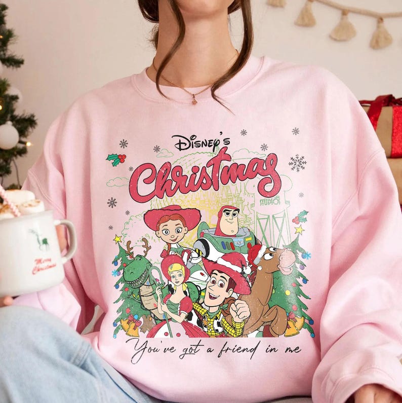 Może przedstawiać: R&oacute;żowa bluza z nadrukiem świątecznym z postaciami z filmu Disneya. Na bluzie znajdują się postacie z Toy Story, takie jak Woody i Buzz Lightyear. Widoczne są napisy "Disney's Christmas" i "You've got a friend in me".