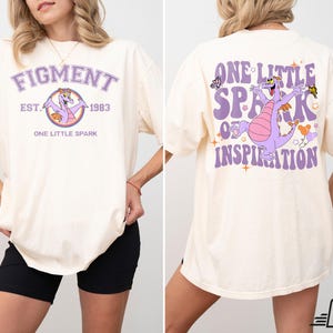 Op de afbeelding: Een licht beige T-shirt met een paarse draak afbeelding en de tekst "Figment Est. 1983 One Little Spark" op de voorkant. De achterkant van het shirt heeft een paarse draak afbeelding en de tekst "One Little Spark of Inspiration".