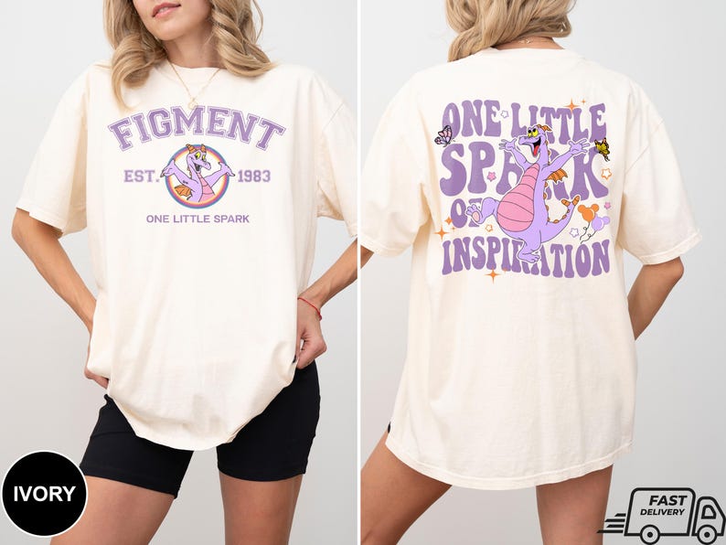 Op de afbeelding: Een licht beige T-shirt met een paarse draak afbeelding en de tekst "Figment Est. 1983 One Little Spark" op de voorkant. De achterkant van het shirt heeft een paarse draak afbeelding en de tekst "One Little Spark of Inspiration".