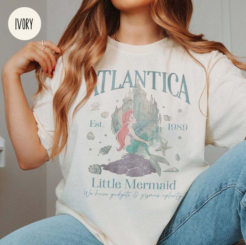 May include: A white t-shirt with a vintage-inspired graphic of Ariel from The Little Mermaid. The graphic features the words "Atlantica" and "Little Mermaid" along with the text "Est. 1989".