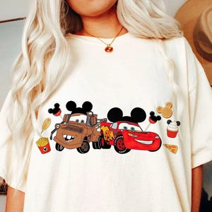Puede incluir: Camiseta color crema con un gr&aacute;fico de dos coches de dibujos animados, uno marr&oacute;n y otro rojo, con orejas de Mickey Mouse. El dise&ntilde;o incluye iconos de comida como patatas fritas, un gofre y una magdalena. La camiseta es de estilo casual.
